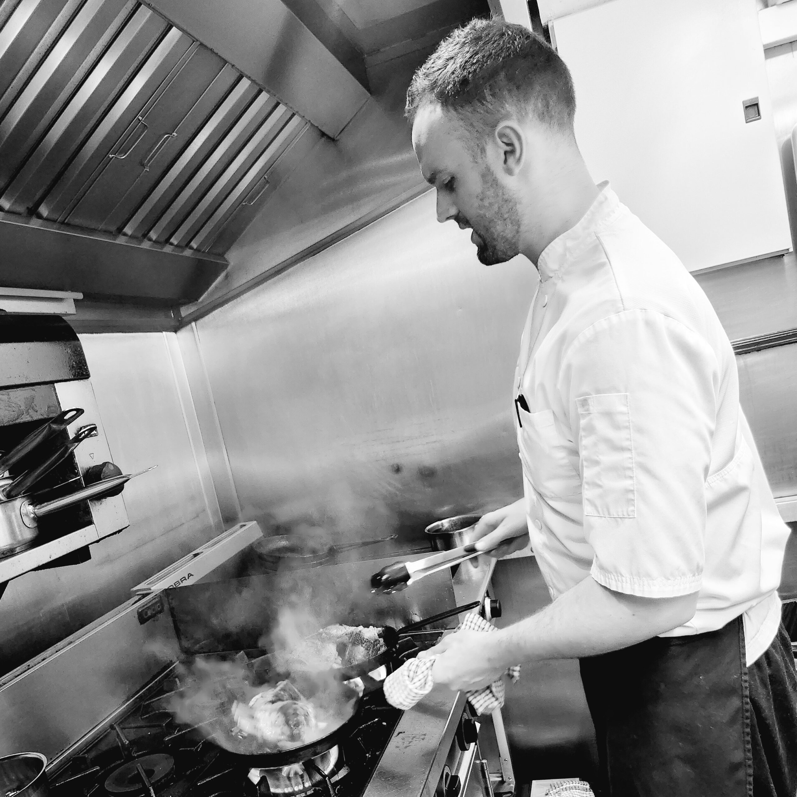 Marek Ivasienko in chef whites, holding a smoking pan over an oven hob.